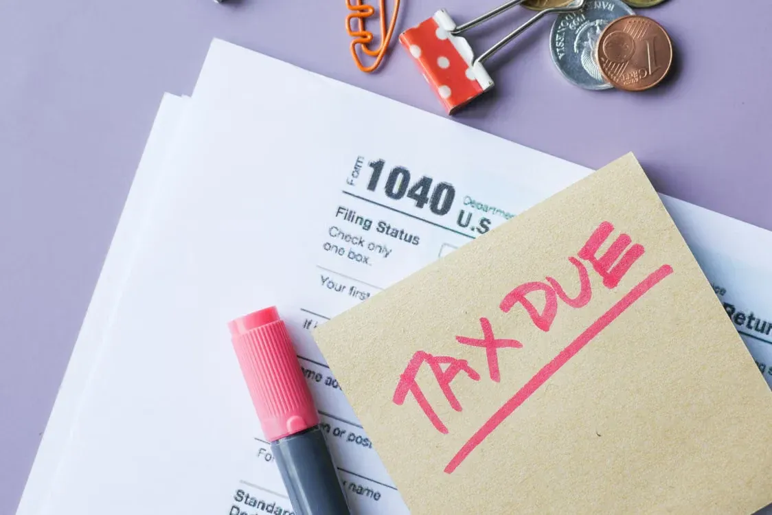 Form 1040 tax papers with a sticky note reading Tax Due, a pink marker, coins and binder clips